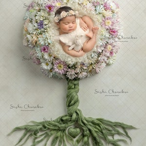 May include: A newborn baby sleeping in a nest of white and pink flowers. The nest is attached to a green yarn tree trunk with roots spreading out on a white background.