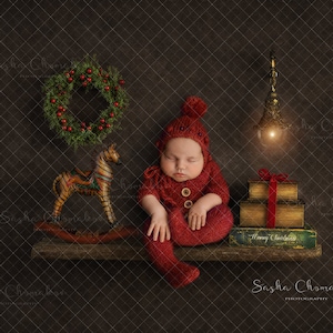 Może przedstawiać: Śpiące dziecko w czerwonym dzianinowym ubranku siedzi na drewnianej półce z konikiem na biegunach, wieńcem i stosem książek. Półka jest ozdobiona girlandą światełek i tabliczką z napisem "Wesołych Świąt".