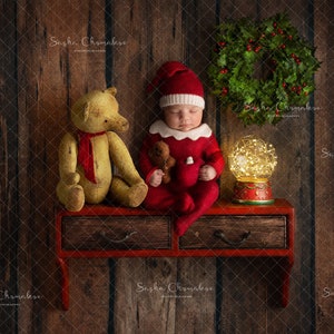 Puede incluir: Un bebé dormido con un traje de Papá Noel rojo y blanco se sienta en un estante rojo con un oso de peluche y una bola de nieve con luces. El estante está contra un fondo de madera con una corona verde colgada en la pared.