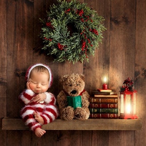 May include: A sleeping baby wearing a red and white striped knitted outfit sits on a wooden shelf with a teddy bear holding a book and a lit candle next to a lantern. A Christmas wreath hangs on the wall behind the shelf.