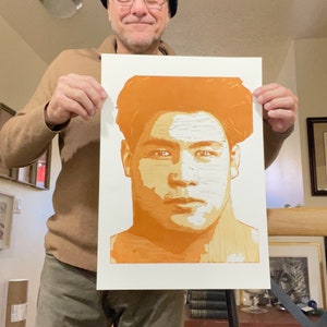 May include: A person holds up a print of a man's face. The print is in shades of orange and white. The man in the print has short, dark hair and a serious expression.
