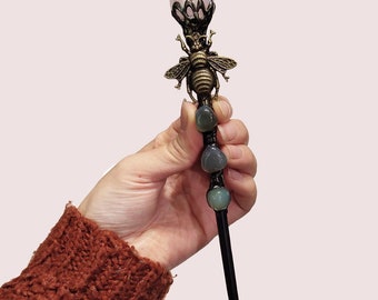 May include: A decorative hair stick featuring a bronze bee, three green stones, and a pink crystal. The hair accessory has a black wooden stick and is held against a light pink background.
