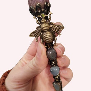 May include: A decorative hair stick with a bronze bee motif, a large pink gemstone, and three gray heart-shaped stones. The stick is held in a hand. The top gemstone is approximately 2.5 cm in diameter.