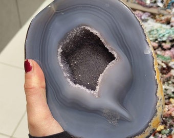 May include: A large, gray agate geode with a hollow center lined with sparkling crystals. The geode is being held in a hand.