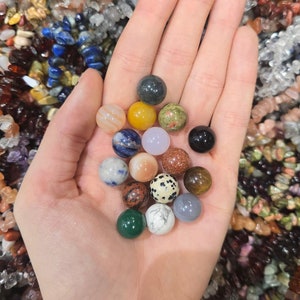 May include: A hand holding a variety of small, round, polished stones in different colours and patterns. The stones are likely gemstones or crystals.