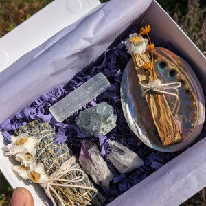 May include: A white box containing purple shredded paper, a selenite wand, a blue-green crystal, a bundle of dried lavender with white flowers, amethyst and clear quartz crystals, a wooden stick, and an abalone shell.