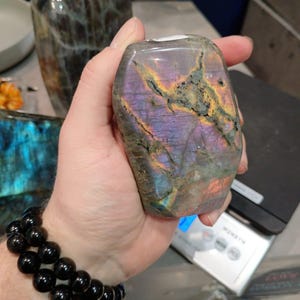 Puede incluir: Una mano sostiene una gran gema de labradorita iridiscente, de color p&uacute;rpura y gris. La piedra tiene un patr&oacute;n &uacute;nico y natural de vetas y remolinos. La piedra se est&aacute; pesando en una b&aacute;scula digital.