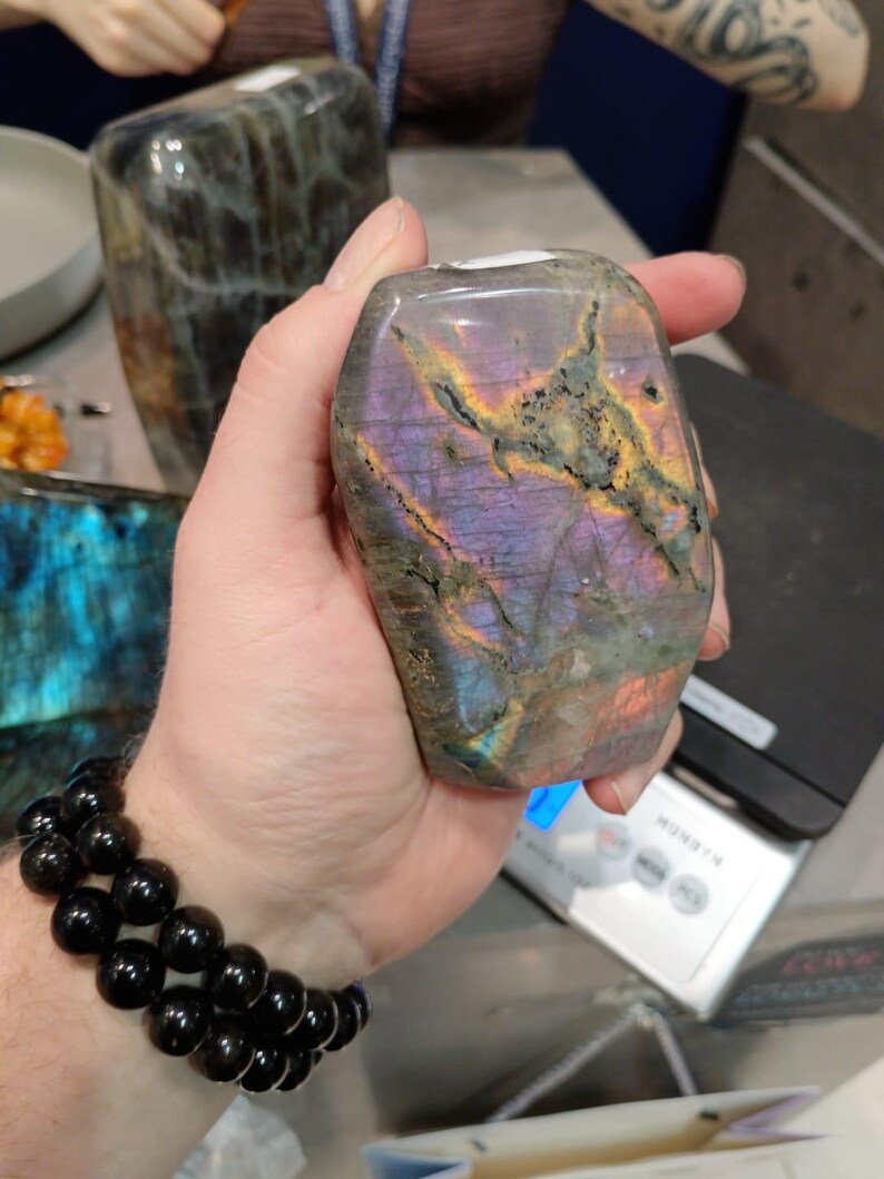 Puede incluir: Una mano sostiene una gran gema de labradorita iridiscente, de color p&uacute;rpura y gris. La piedra tiene un patr&oacute;n &uacute;nico y natural de vetas y remolinos. La piedra se est&aacute; pesando en una b&aacute;scula digital.