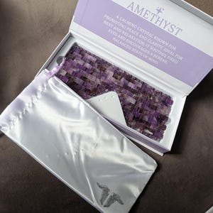 May include: An amethyst eye mask in a white box. The box has the word "AMETHYST" printed on it. The mask is made of small, purple amethyst stones. A white satin pouch with a wing design is included.