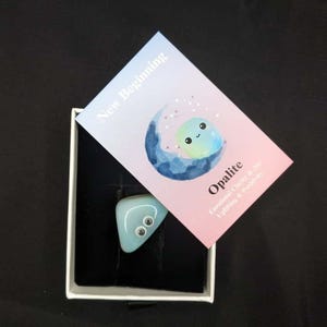 Puede incluir: Un colgante de opalita azul claro con ojos saltones descansa en una caja blanca. Una tarjeta con las palabras "New Beginning" y "Opalite" está colocada encima. La tarjeta tiene una ilustración de acuarela de una luna y una cara sonriente.