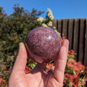 May include: A large, polished, purple amethyst sphere with a cloudy, translucent appearance. The sphere is being held in a hand.