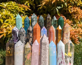Op de afbeelding: Een verzameling van 25 gepolijste kristallen in verschillende kleuren en texturen, waaronder helder, roze, groen, bruin en wit. De kristallen zijn in een rij op een oppervlak gerangschikt.