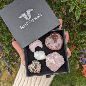 Op de afbeelding: Een zwarte doos met de tekst "SpiritCrystals" op de deksel, die een verzameling roze en zwarte hartvormige kristallen, een halve maan kristal, een wit kristal en een groot roze kristal bevat.