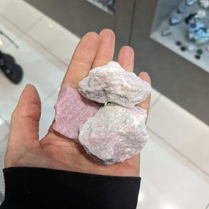 May include: Three pink and white rough cut stones in a hand. The stones are a light pink color with white streaks and veins.