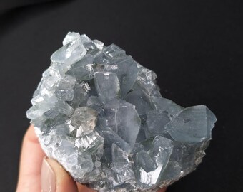 May include: A cluster of pale blue celestite crystals. The crystals have a translucent appearance and are arranged in a jagged, irregular formation. The base of the cluster is a rough, off-white color. The background is black.