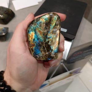 Puede incluir: Una gran gema de labradorita pulida e iridiscente con tonos azules, verdes y dorados. La piedra se sostiene en una mano.