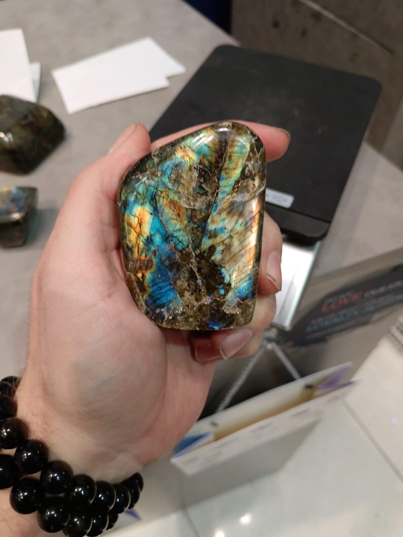 Puede incluir: Una gran gema de labradorita pulida e iridiscente con tonos azules, verdes y dorados. La piedra se sostiene en una mano.