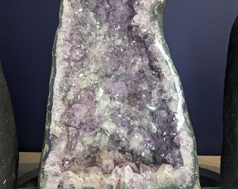 May include: A large amethyst geode with a dark gray base. The interior is filled with sparkling purple crystals. The geode has a rough, natural exterior and a label with the numbers "185" and "116".