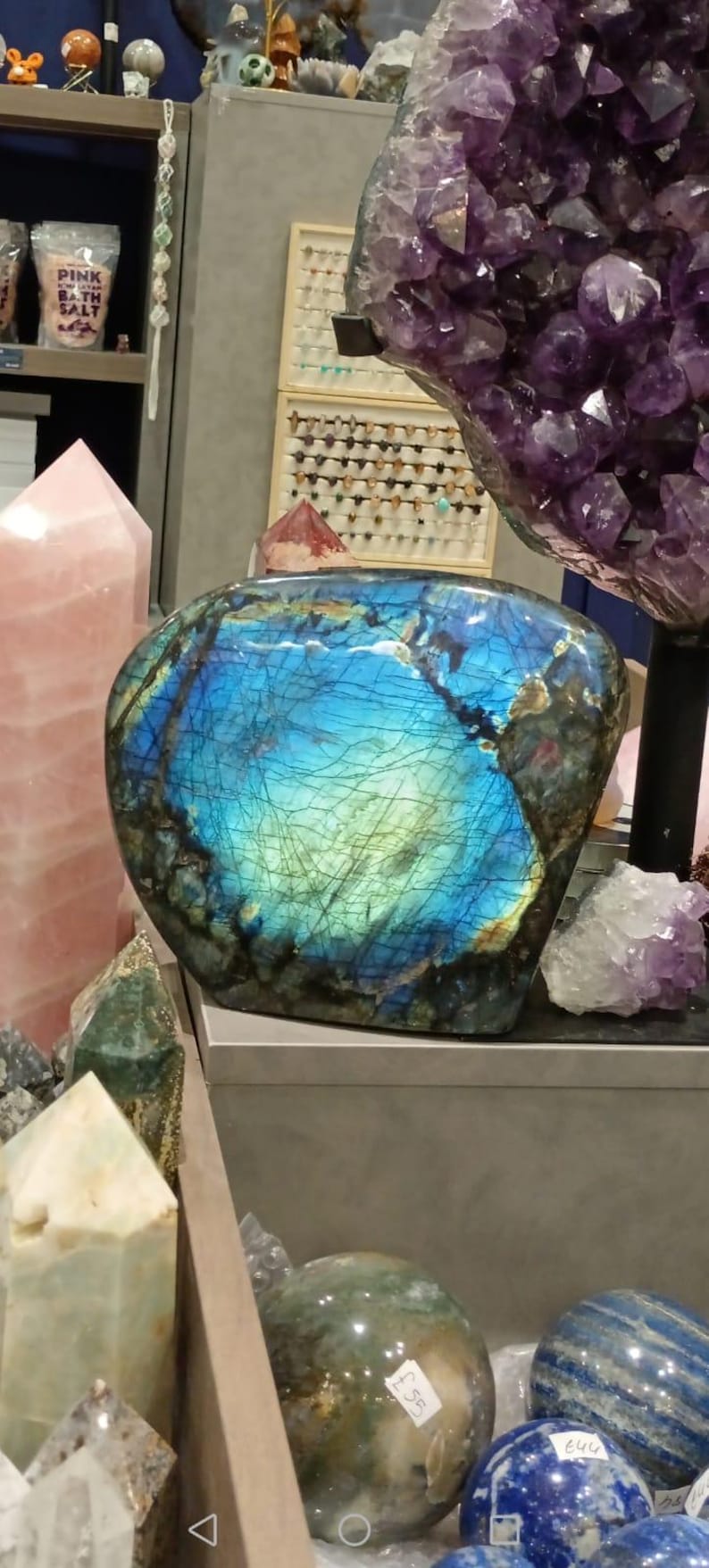 Puede incluir: Un gran cristal de labradorita azul iridiscente con una superficie lisa y redondeada. El cristal se exhibe en un estante con otros cristales, incluyendo un cuarzo rosa y una amatista p&uacute;rpura.