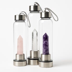 Può includere: Tre bottiglie d'acqua di vetro con tappi e cinturini argentati. Ogni bottiglia contiene un cristallo: un quarzo rosa, un quarzo trasparente e un'ametista viola.