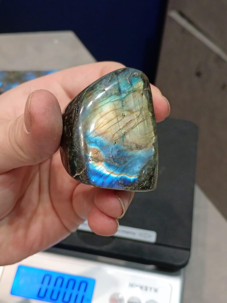 Puede incluir: Una piedra preciosa de labradorita azul y verde con destellos iridiscentes. La piedra preciosa es lisa y pulida, y se est&aacute; sosteniendo en una mano.