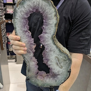Könnte beinhalten: Große Amethyst-Geode mit einem dunkelvioletten Innenraum und einem hellgrauen und grünen Außenrand. Die Geode wird aufrecht gehalten und zeigt die Kristallformationen im Inneren. Die Geode ist auf einem schwarzen Ständer montiert.