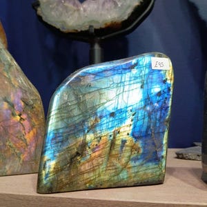 Puede incluir: Un gran cristal de labradorita azul iridiscente con una etiqueta de precio de "&pound;95" adjunta. El cristal tiene una textura rugosa y natural y es una opci&oacute;n popular para coleccionistas y entusiastas de la metaf&iacute;sica.