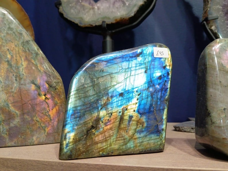 Puede incluir: Un gran cristal de labradorita azul iridiscente con una etiqueta de precio de "&pound;95" adjunta. El cristal tiene una textura rugosa y natural y es una opci&oacute;n popular para coleccionistas y entusiastas de la metaf&iacute;sica.
