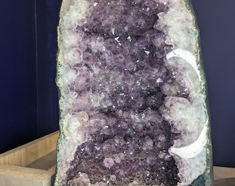 May include: Large amethyst geode with a dark purple interior and white crystalline edges. The geode is mounted on a black base, with a price tag visible. The interior is filled with sparkling crystal formations.