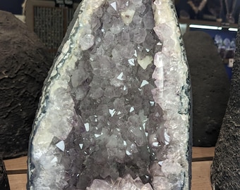 May include: A large amethyst geode with a dark exterior and a cavity filled with sparkling purple crystals. The geode is displayed on a wooden shelf, showcasing its natural beauty and geological formation.