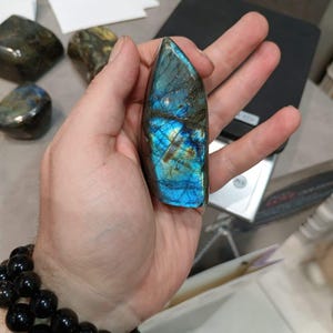 Puede incluir: Un gran cristal de labradorita azul y verde iridiscente con una punta puntiaguda. El cristal est&aacute; siendo sostenido en una mano.