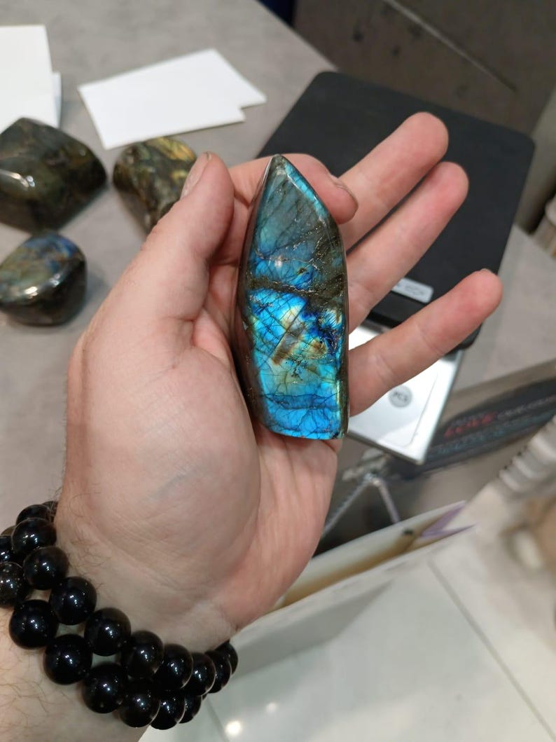 Puede incluir: Un gran cristal de labradorita azul y verde iridiscente con una punta puntiaguda. El cristal est&aacute; siendo sostenido en una mano.