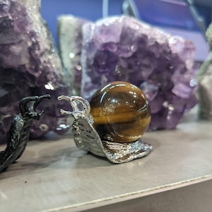 Könnte beinhalten: Eine dekorative Schneckenfigur mit einer polierten Tigeraugenkugel als Gehäuse. Der Schneckenkörper ist silberfarben, und die Kugel hat braune und goldene Bänder. Im Hintergrund befinden sich Amethystkristalle.