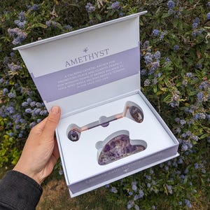 May include: A white box with a purple interior holds an amethyst face roller and a gua sha tool. The box has the word "AMETHYST" and text that reads "I choose calm in every moment." The roller and tool are purple and rose gold.
