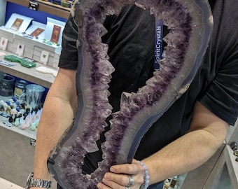 May include: A large amethyst geode with a natural opening, displaying a purple and white crystal formation. The geode is on a black stand and is being held by a person. The text "SpiritCrystals" is visible on the geode.