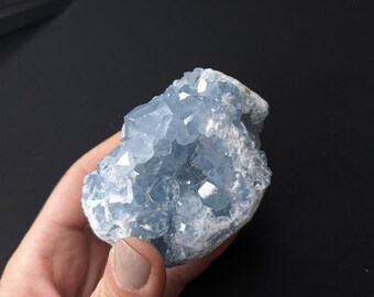 May include: A light blue celestite crystal cluster. The crystal has a geode-like structure with multiple facets and a rough, textured surface. The color is a soft, pale blue with some areas of white. The crystal is held in a hand against a black background.