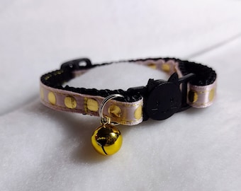 May include: A pet collar featuring a light pink ribbon with gold polka dots layered over a black nylon strap. The collar includes a black plastic buckle shaped like a cat face and a small, round, shiny gold-colored metal bell attached to a ring.