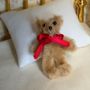 May include: A miniature teddy bear with tan fur and a bright red ribbon tied in a bow around its neck. The small bear is resting on a white pillow, suggesting a cozy, comforting theme. The bear's small size and soft appearance make it a charming collectible.