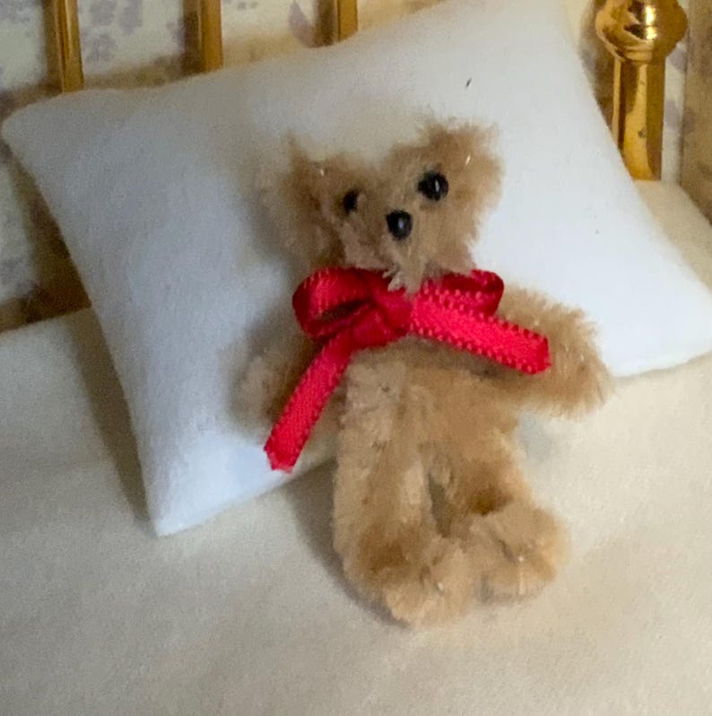 May include: A miniature teddy bear with tan fur and a bright red ribbon tied in a bow around its neck. The small bear is resting on a white pillow, suggesting a cozy, comforting theme. The bear's small size and soft appearance make it a charming collectible.