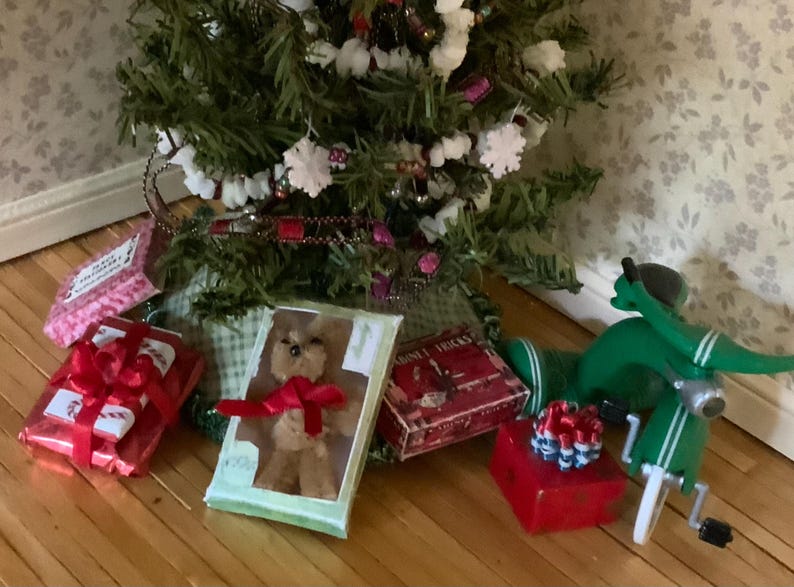May include: A Christmas tree with white snowflake ornaments and red ribbon. Presents wrapped in red paper with red bows are scattered around the base of the tree. A green toy scooter is also present.