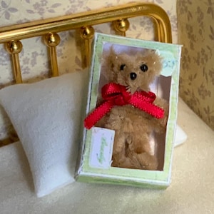 May include: A miniature teddy bear with a red ribbon in a green and white box. The bear is light brown and has black eyes. The box is rectangular and has a clear plastic front. The scene includes a white pillow and a gold bed frame.