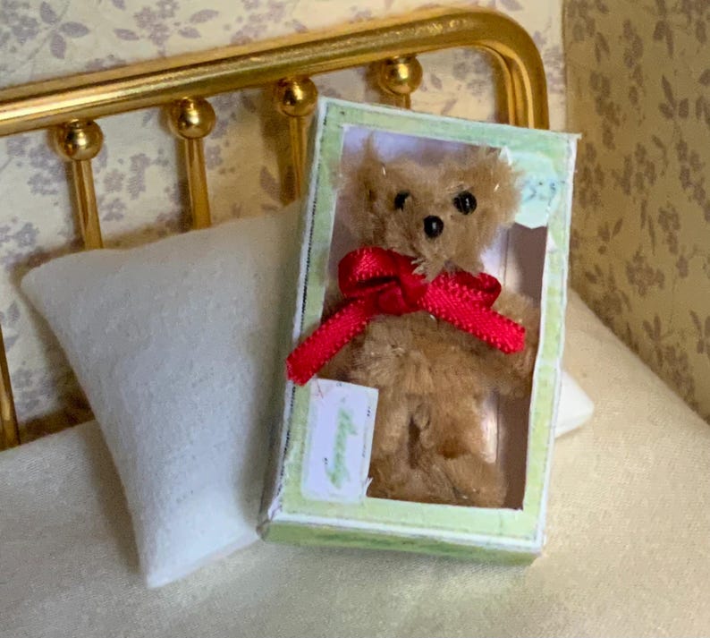 May include: A miniature teddy bear with a red ribbon in a green and white box. The bear is light brown and has black eyes. The box is rectangular and has a clear plastic front. The scene includes a white pillow and a gold bed frame.