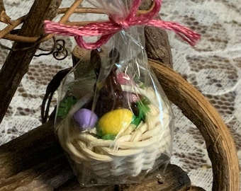 May include: A small white wicker basket filled with a chocolate bunny, a yellow and a purple egg, and green leaves. The basket is wrapped in clear cellophane and tied with a pink ribbon.