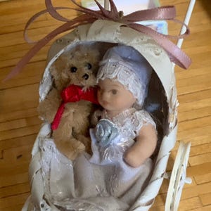 May include: A miniature baby doll in a white dress and blue bonnet sits in a decorative white carriage with a small teddy bear. The teddy bear has a red bow. The carriage has a pink ribbon.