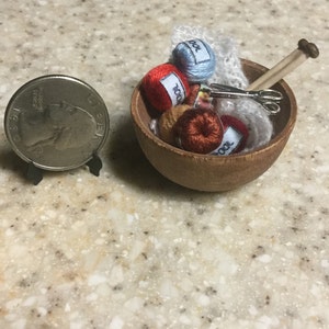 May include: A wooden bowl filled with yarn balls, a pair of scissors, and a knitting needle. The yarn balls are red, orange, and blue. The bowl is on a light brown surface.