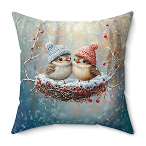 May include: Square throw pillow featuring a winter scene with two birds wearing knitted hats, nestled in a snowy nest with red berries. The background shows a snowy forest with a soft, blurred effect. The pillow has a blue and white color scheme.