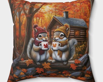 Könnte beinhalten: Quadratisches Kissen mit zwei Cartoon-Eichhörnchen, die Mützen und Schals tragen und Tassen mit Ahornblatt-Designs halten. Die Eichhörnchen stehen vor einer Hütte in einer Herbstwaldkulisse mit orangefarbenen und roten Blättern.
