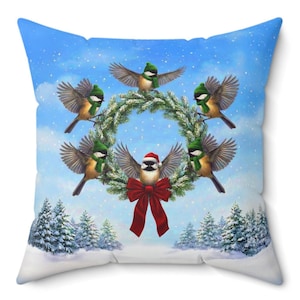 May include: A square cushion with a winter design. It depicts six birds around a wreath, wearing green hats and scarves. One bird has a Santa hat and red bow. The background shows a snowy landscape and a blue sky with falling snow.