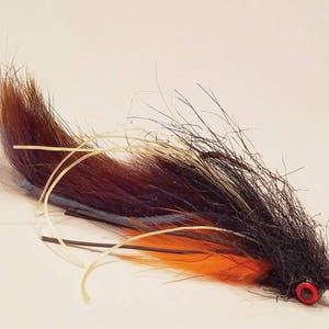 Puede incluir: Un señuelo de pesca con mosca negro, marrón y naranja con un ojo rojo.