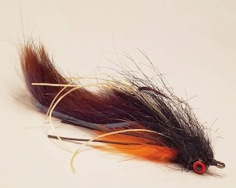 Può includere: Un'esca da pesca a mosca nera, marrone e arancione con un occhio rosso.